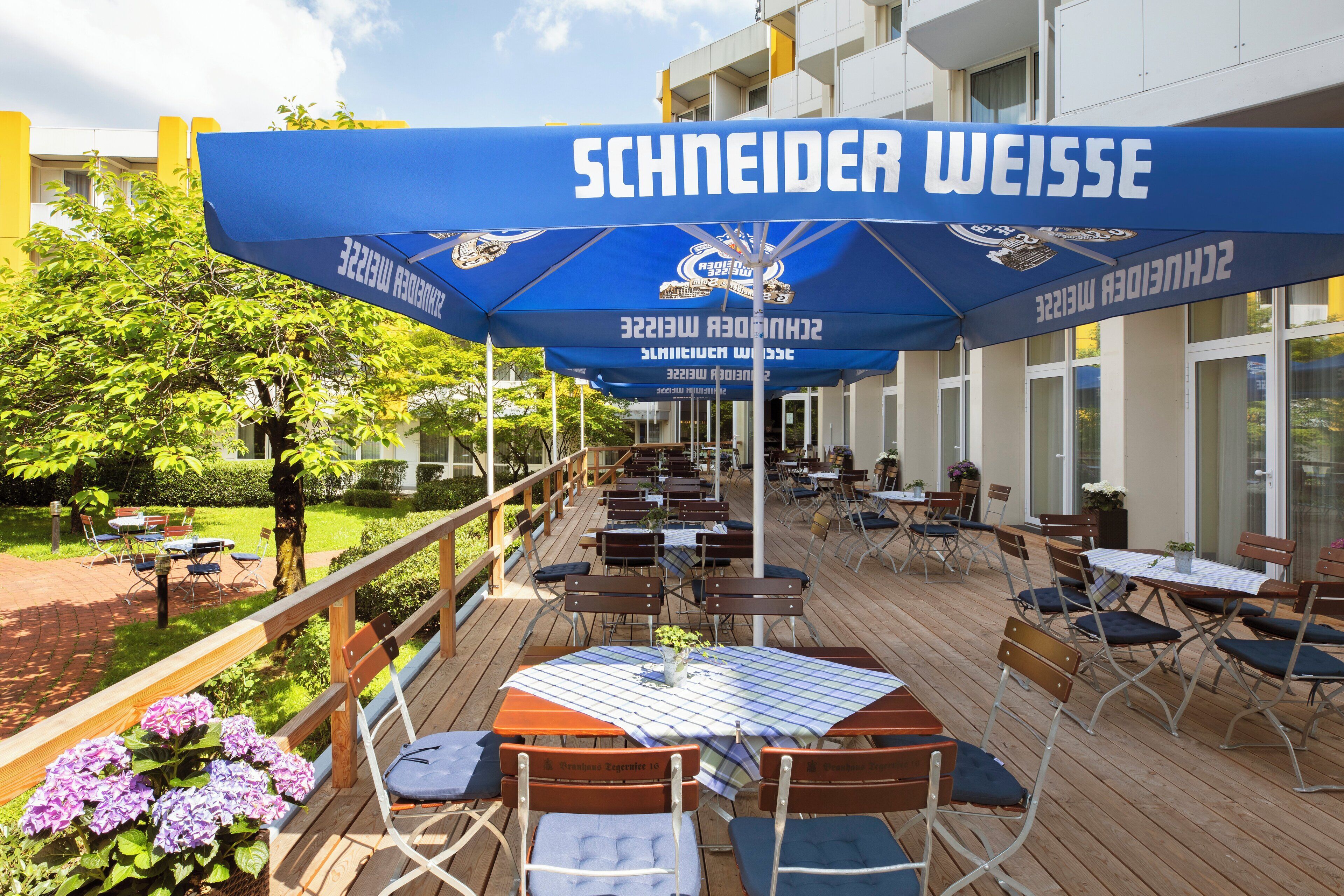 An outdoor seating area with tables and chairs, covered by blue umbrellas with 'SCHNEIDER WEISSE' branding, and a wooden deck.