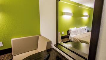 A hotel room with a green wall, a bed with white bedding, a small table with a remote control, and a mirror reflecting the room.