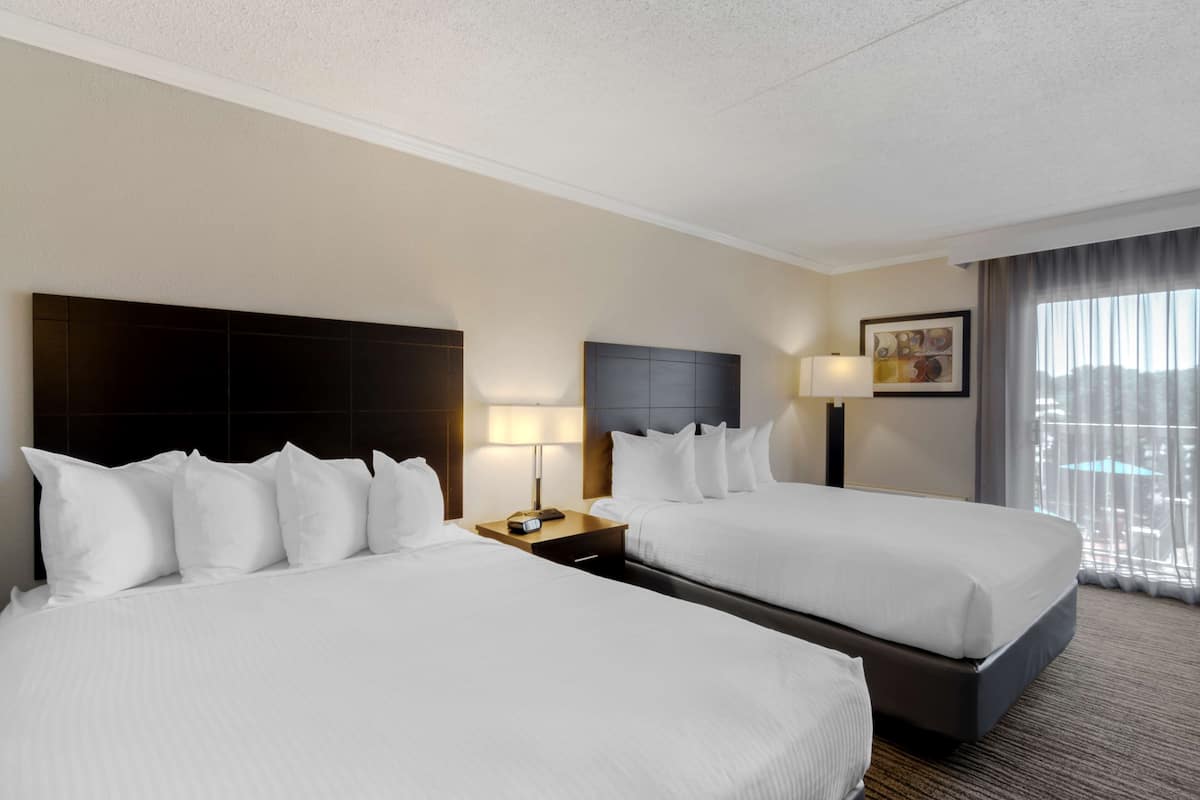 A hotel room with two beds, a nightstand with a lamp, a painting on the wall, and a window with curtains.