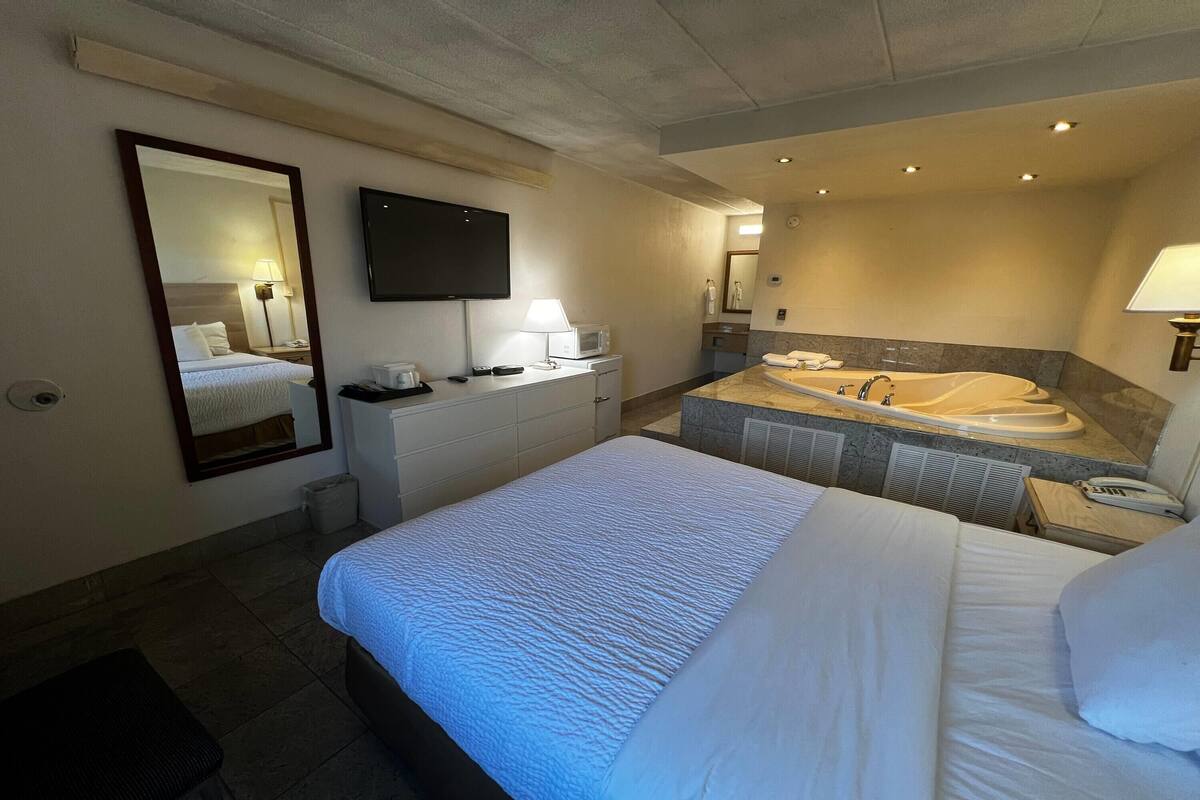 A hotel room with a large bed, a separate bathroom with a bathtub, a flat-screen TV, and a desk with a lamp.