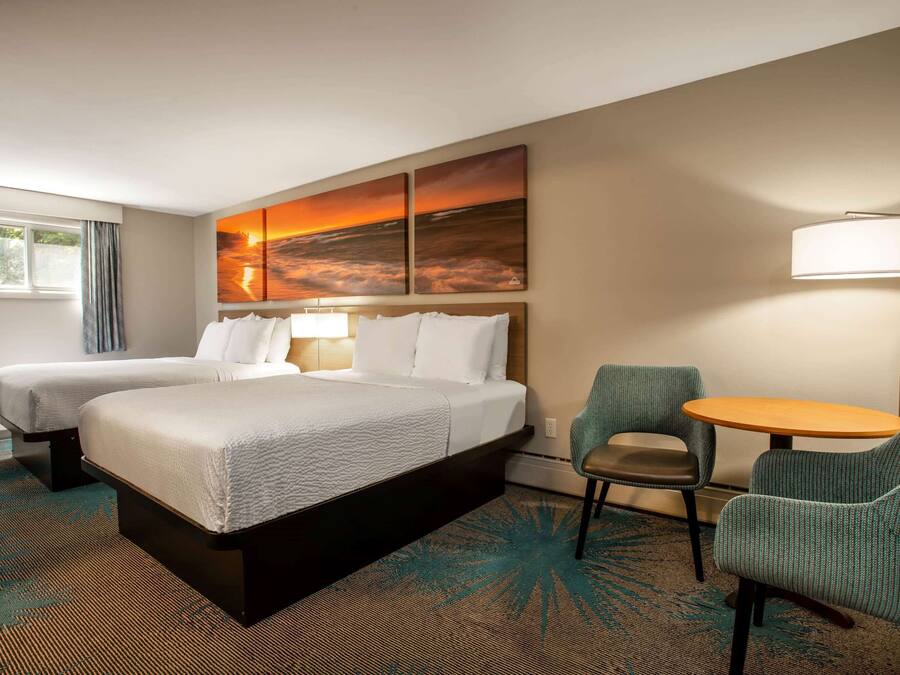 Habitación de hotel con dos camas, un escritorio, una silla, una lámpara y un cuadro de atardecer en la pared.