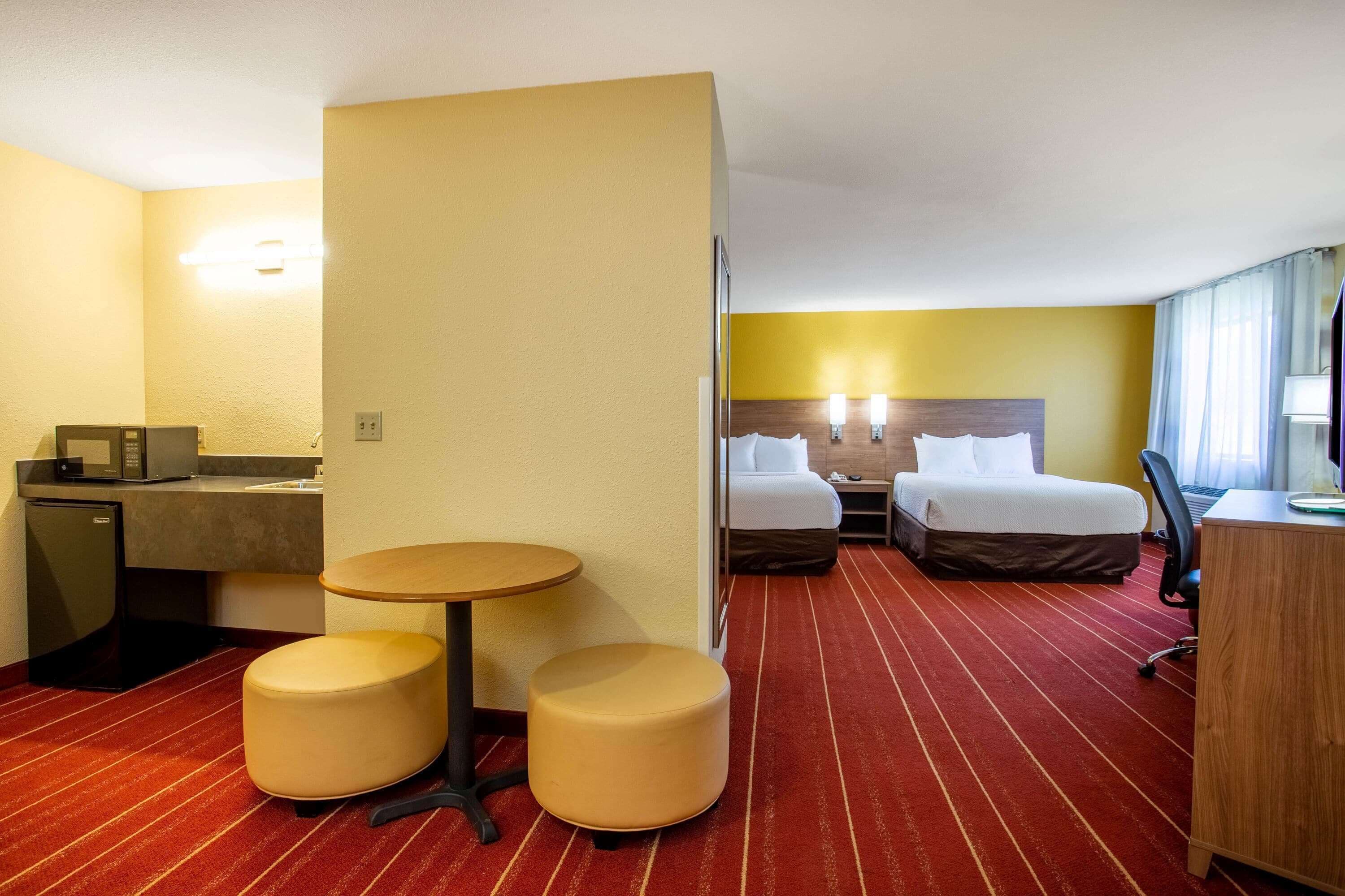 A hotel room with a bed, a desk, a small kitchenette, and two ottomans.