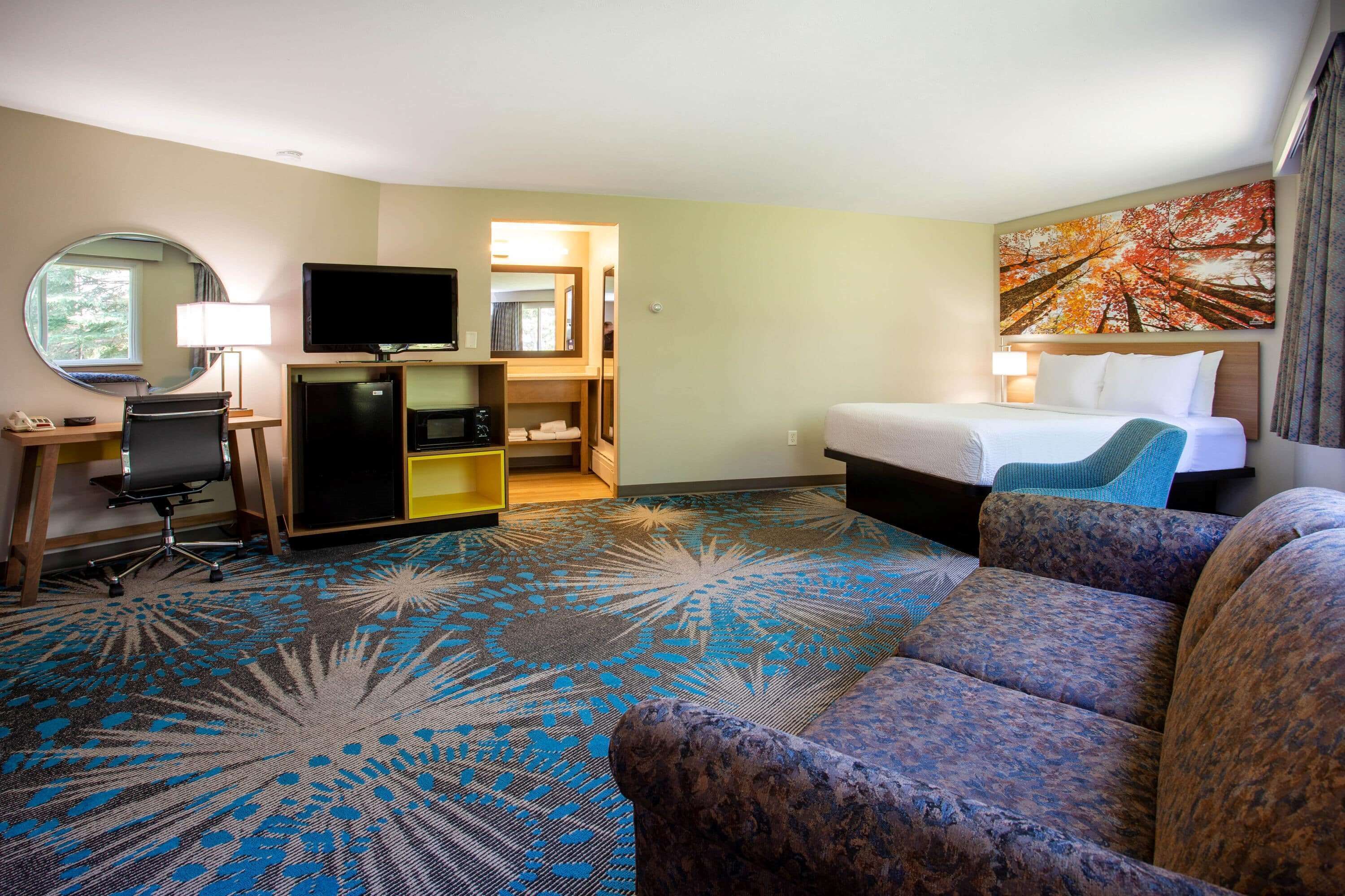 A hotel room with a bed, a sofa, a desk with a chair, a television, a mirror, and a painting on the wall.
