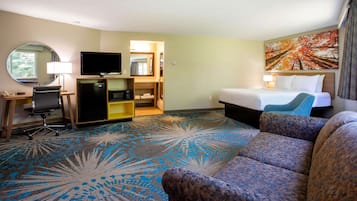A hotel room with a bed, a sofa, a desk with a chair, a television, a mirror, and a painting on the wall.