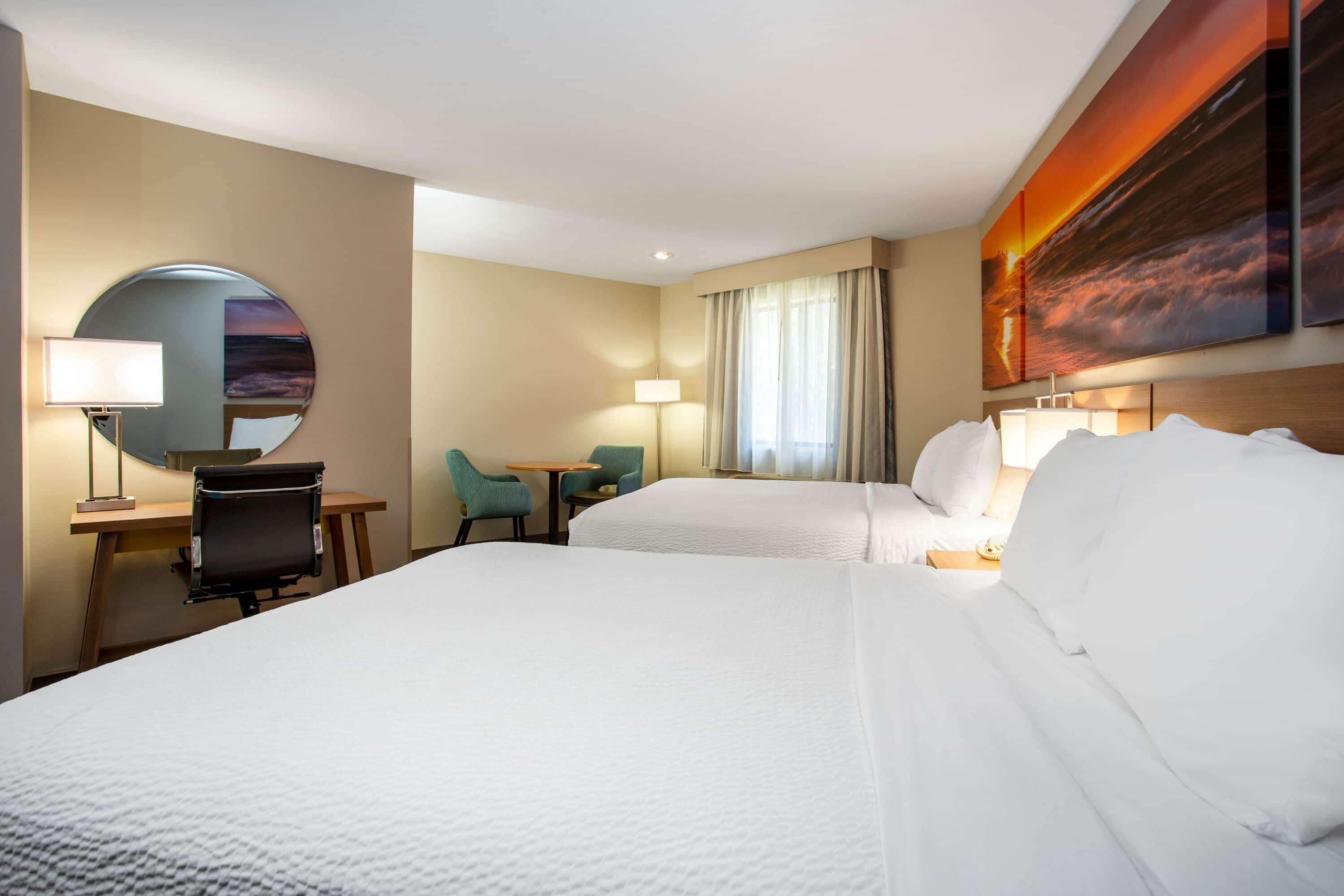 A hotel room with two beds, a desk, a chair, and a round mirror.