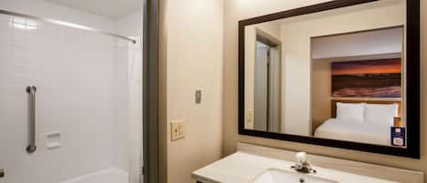 A hotel bathroom with a bathtub, toilet, sink, and a large mirror reflecting a room with a bed and a sunset view.