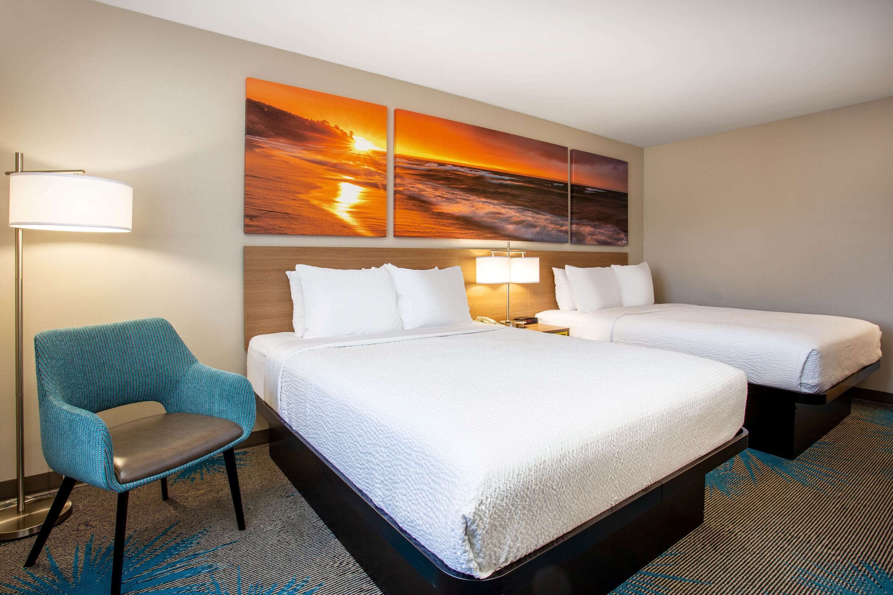 A hotel room with two beds, a blue armchair, a large sunset painting, and a floor lamp.