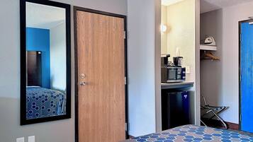 Hotelkamer met een houten deur, een bed met een beddegoed met patroon, een spiegel, een kleine kitchenette met een magnetron en een stoel.