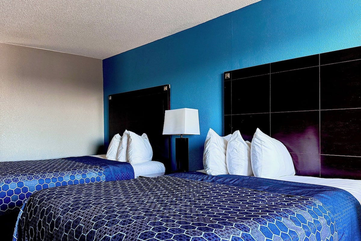 Quarto de hotel com duas camas, colcha azul e branca, cabeceira preta, abajur branco e parede azul.