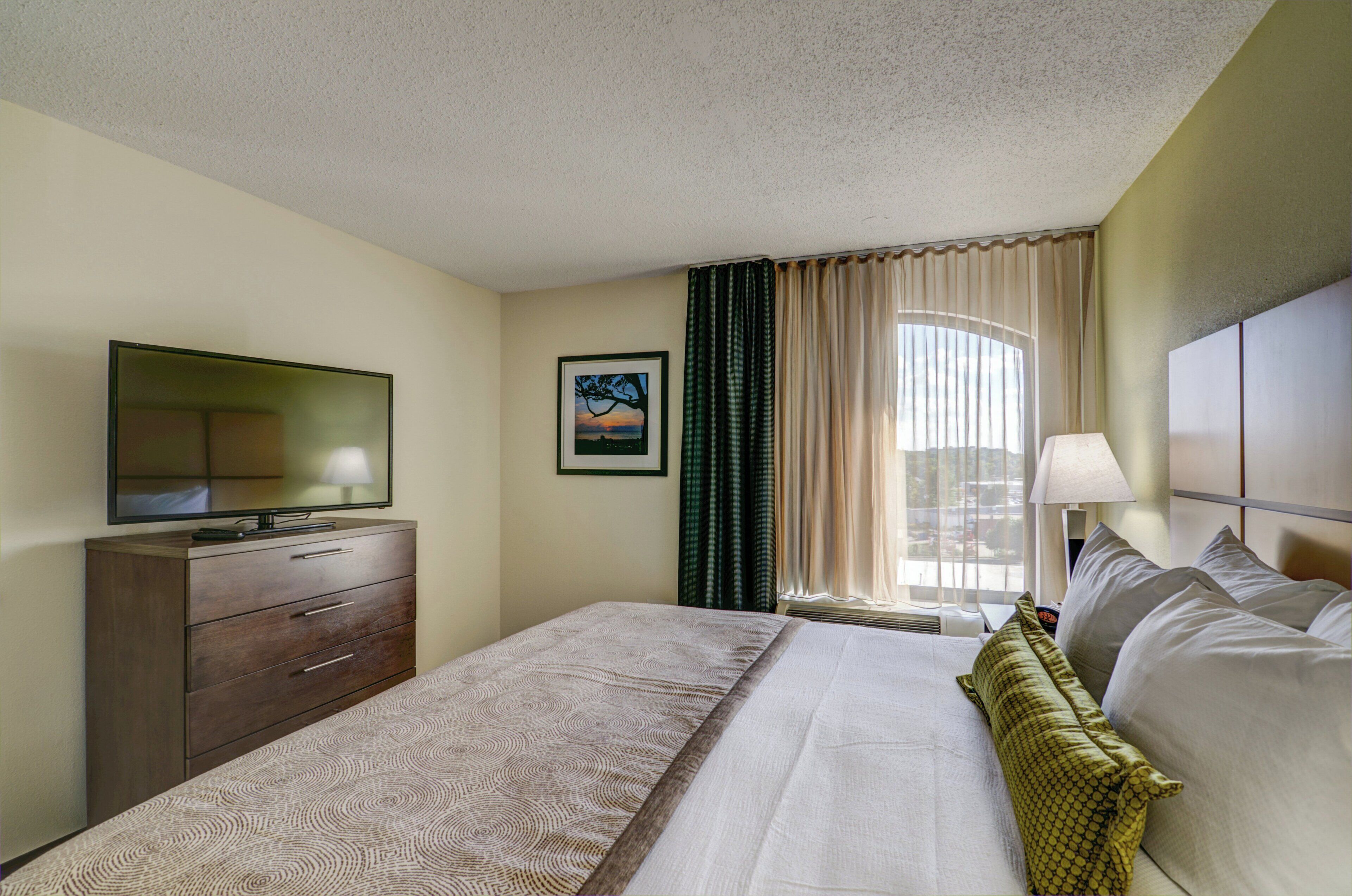 A hotel room with a bed, a TV on a dresser, a window with curtains, and a painting on the wall.