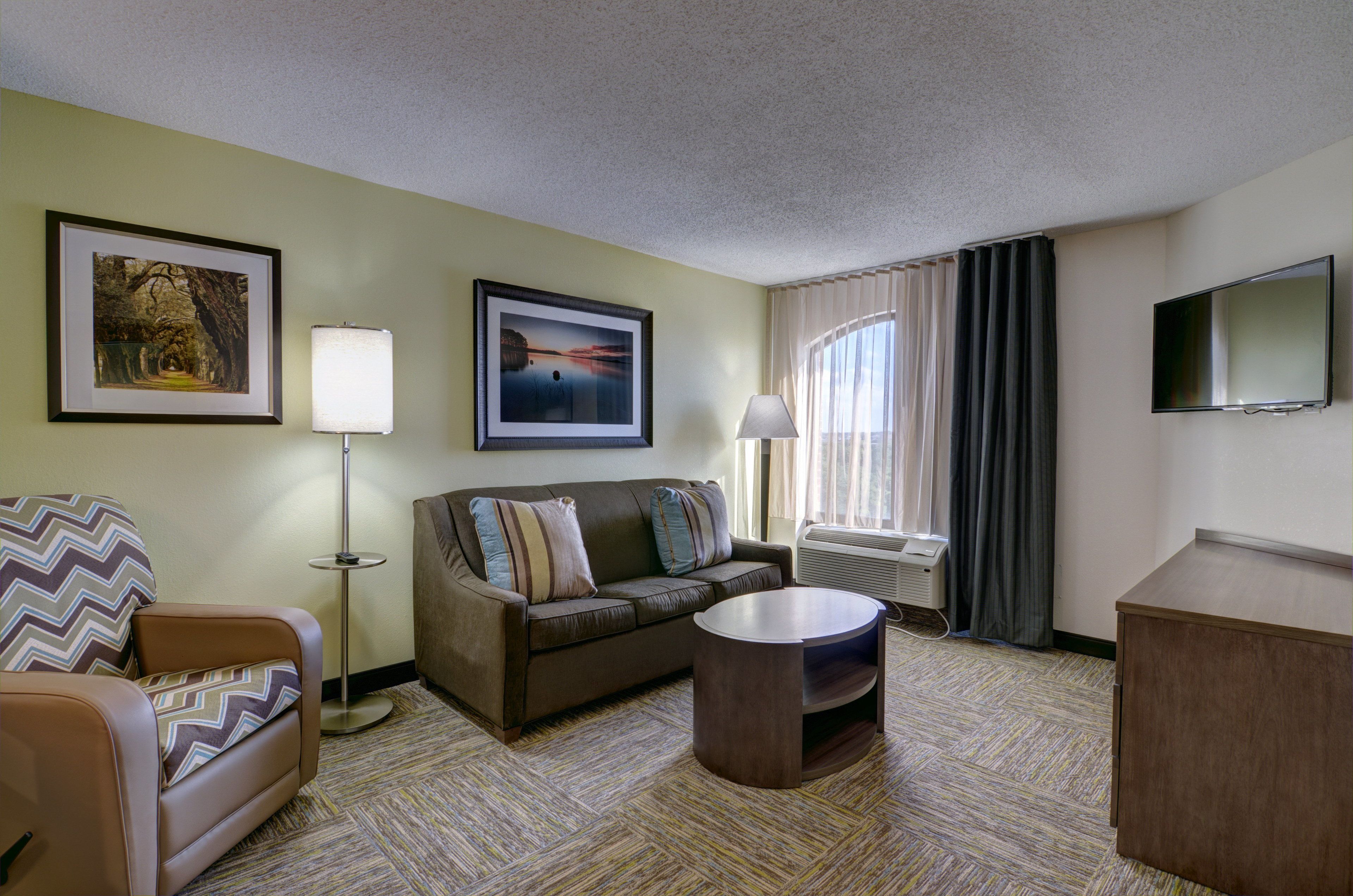 A hotel room with a sofa, a chair, a round coffee table, a flat-screen TV, and two framed pictures on the wall.