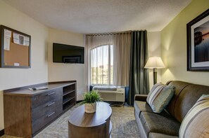 Une chambre d’hôtel dotée d’un téléviseur à écran plat fixé au mur, d’un canapé avec des coussins à rayures, d’une table basse en bois agrémentée d’une plante, d’un bureau avec des tiroirs et d’une fenêtre avec des rideaux.