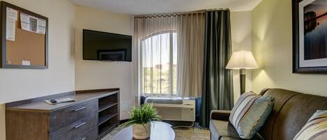 A hotel room with a flat-screen TV mounted on the wall, a sofa, a coffee table, a plant, and a window with curtains.