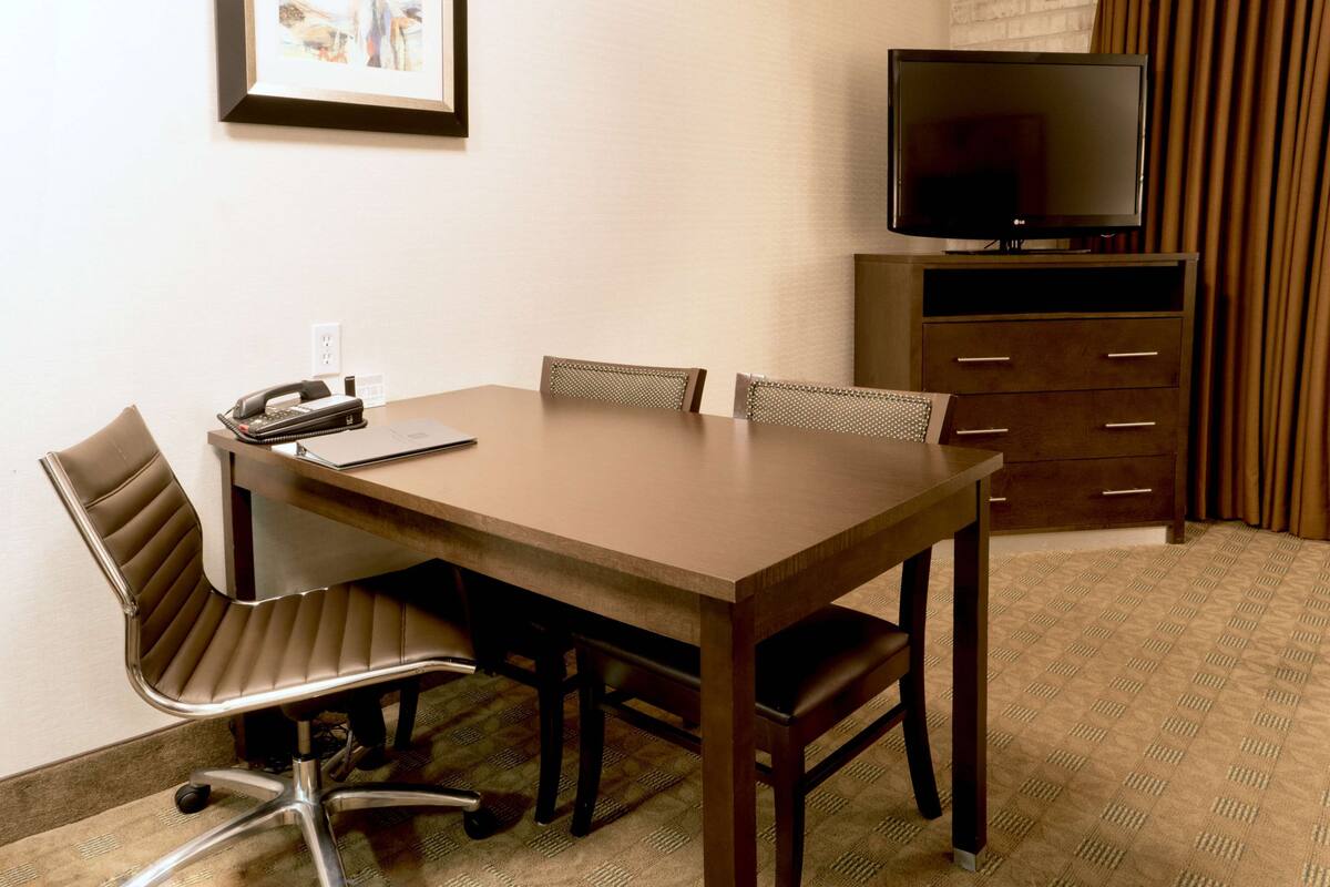 A hotel room with a wooden table, four chairs, a desk with a telephone and a laptop, a television on a stand, and a wardrobe with drawers.