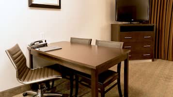 A hotel room with a wooden table, four chairs, a desk with a telephone and a laptop, a television on a stand, and a wardrobe with drawers.