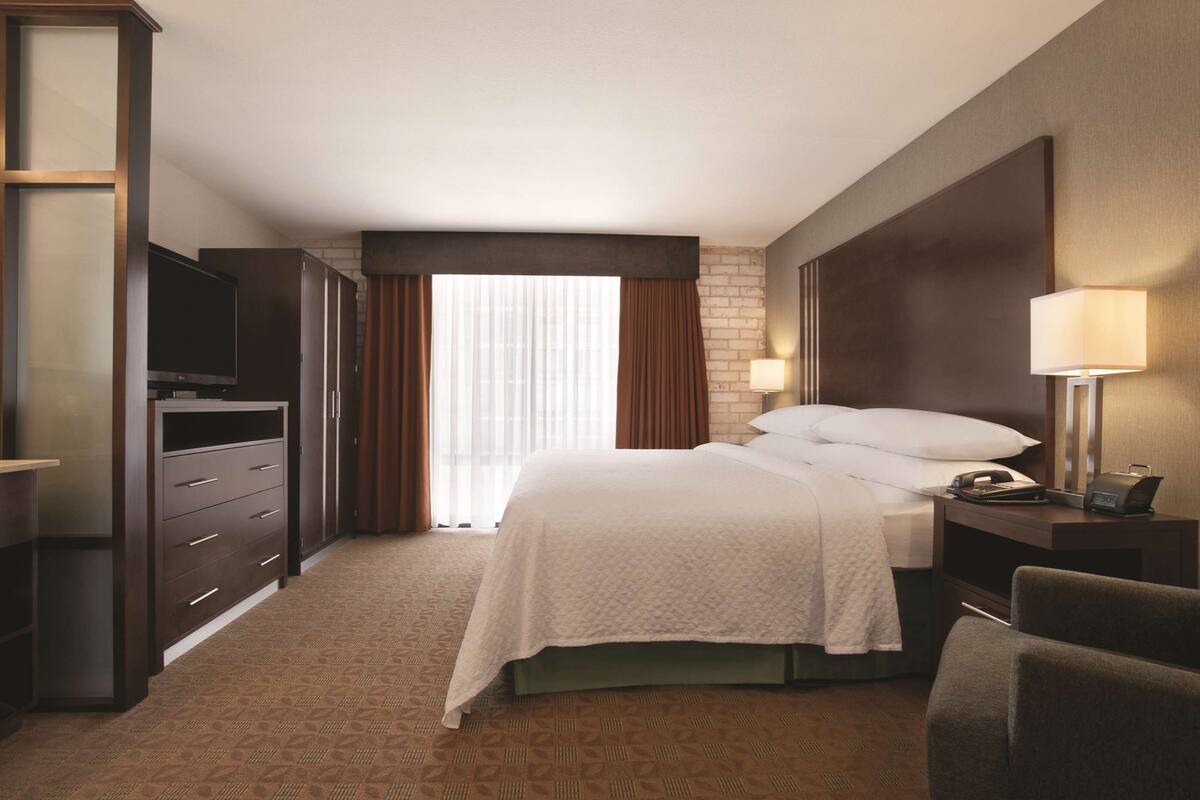A hotel room with a bed, a TV, a desk with a lamp, and a window with curtains.