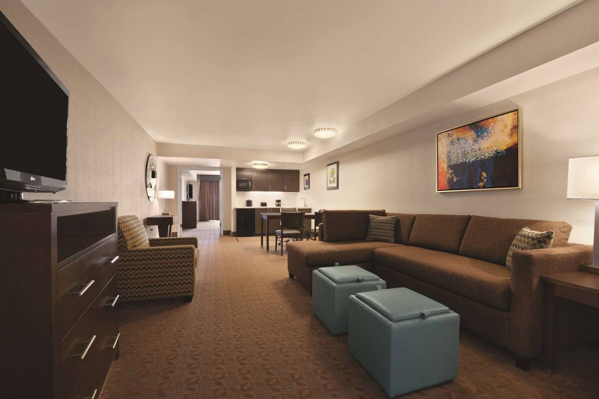 A modern hotel room with a large sectional sofa, a flat-screen TV, a dining area, and a kitchen in the background.
