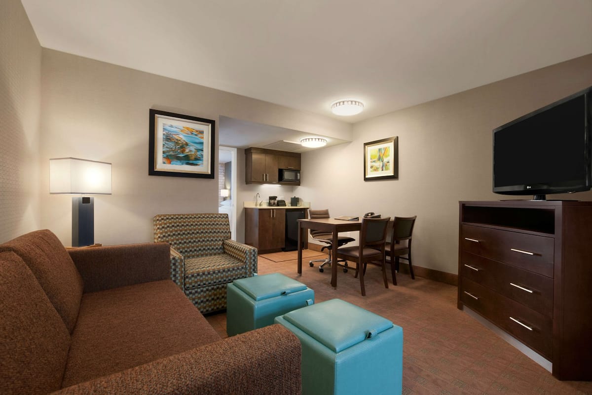 A compact hotel room with a brown sofa, patterned armchair, a dining table with chairs, a kitchenette, a flat-screen TV, and a large cabinet.
