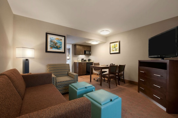 A compact hotel room with a brown sofa, patterned armchair, a dining table with chairs, a kitchenette, a flat-screen TV, and a large cabinet.