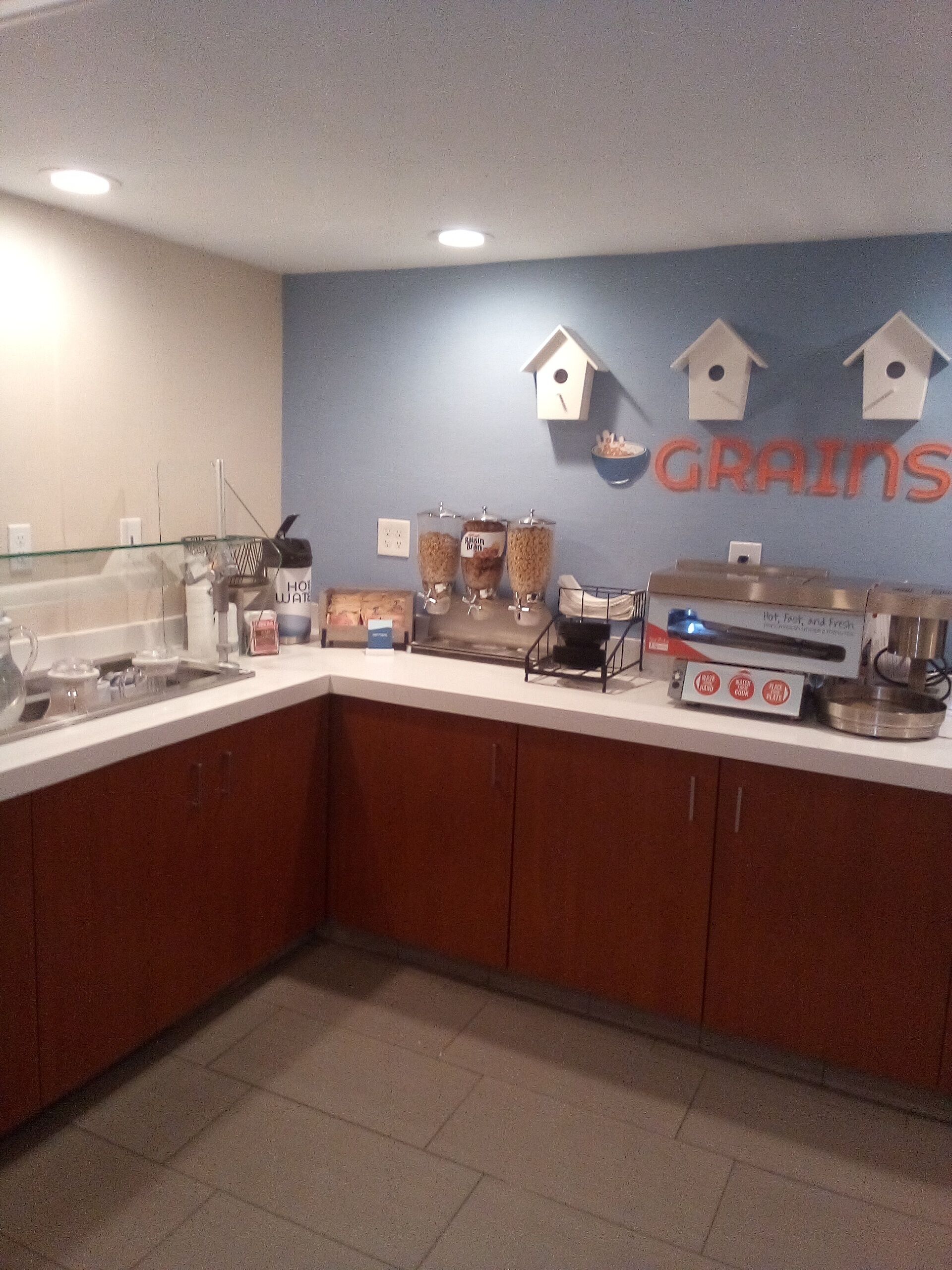 A kitchen area with a counter, food dispensers, a coffee machine, and a wall with a 'Grains' sign.