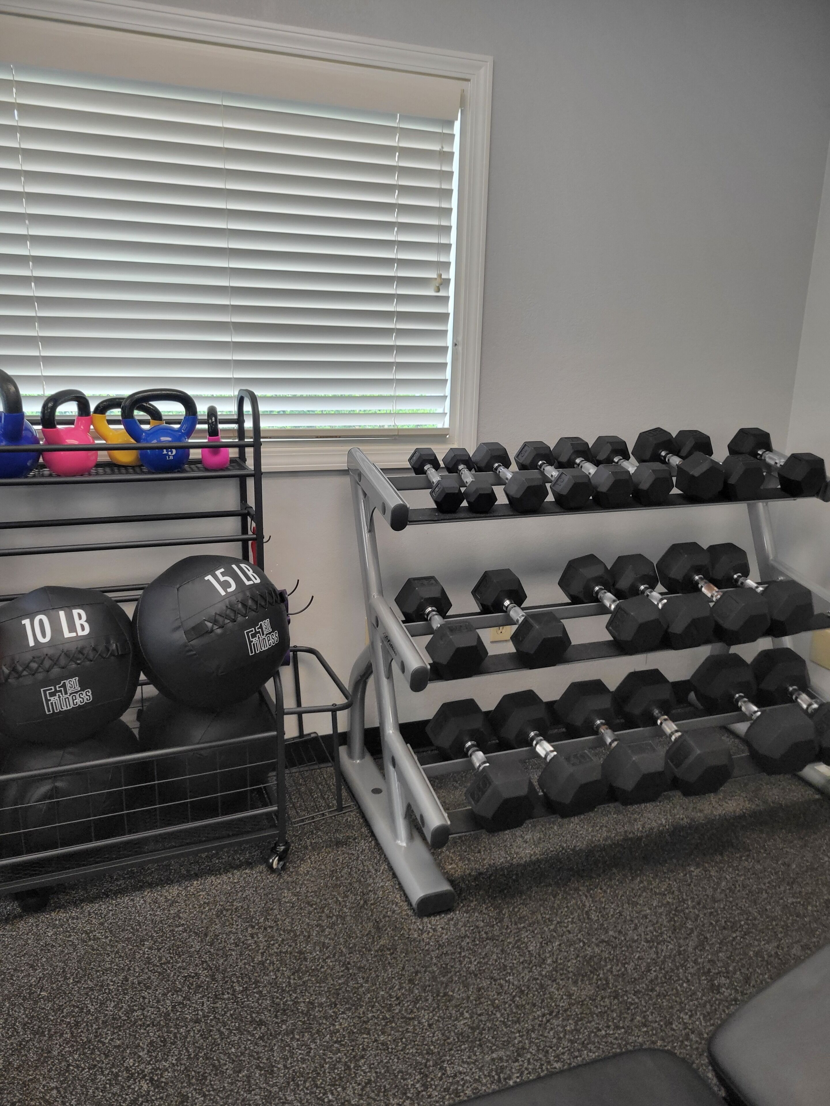 A gym with weightlifting equipment, including kettlebells and dumbbells, and a window with blinds.