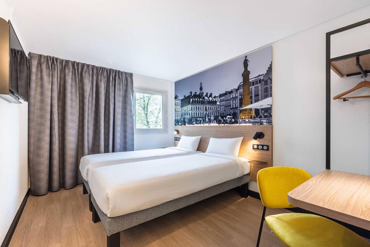 A hotel room with a bed, two chairs, a table, a painting of a cityscape, and a window with curtains.