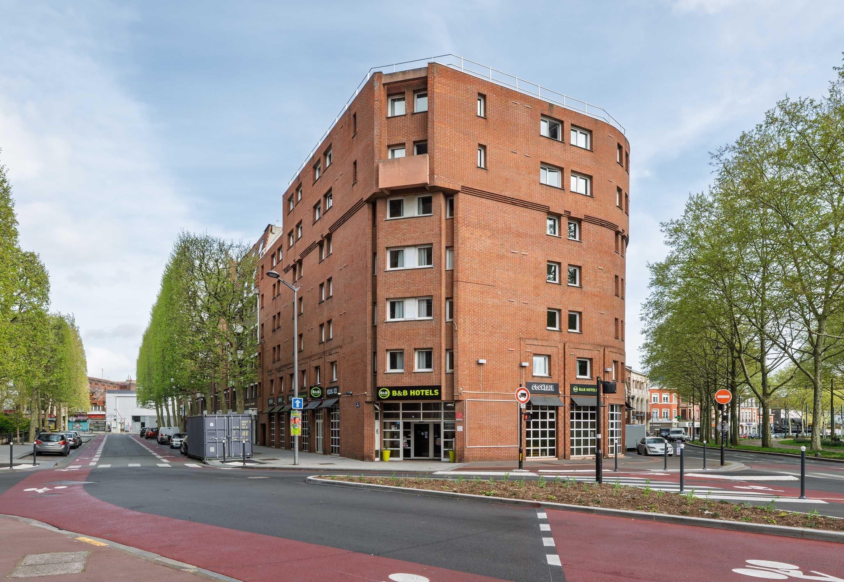 A multi-story hotel with a red brick facade, a ground floor with a visible entrance, and a red bike lane in front.