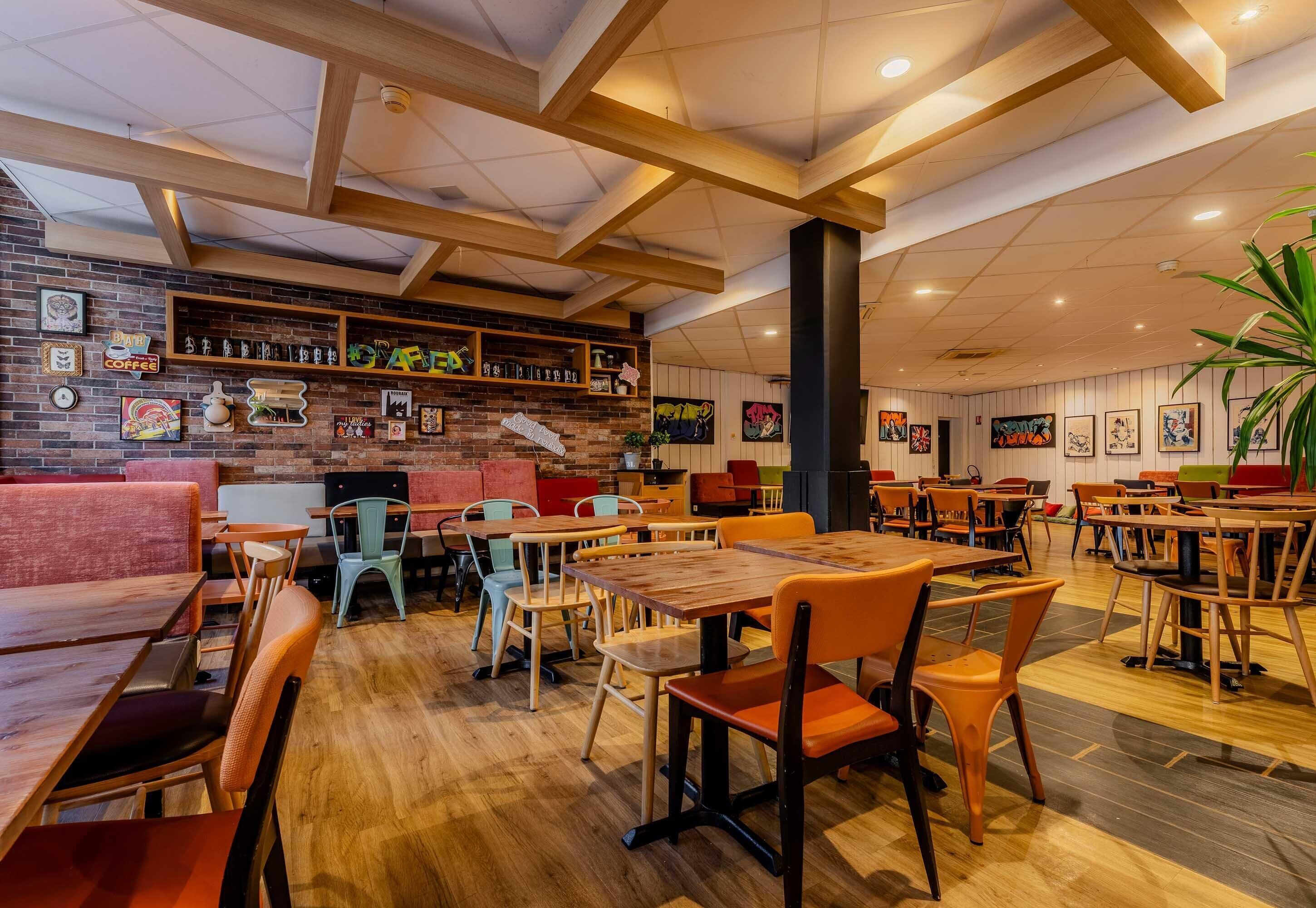En restaurantindretning med træborde og stole, en murstensvæg med kunst og et loft med synlige bjælker.