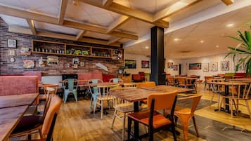 Een restaurantinterieur met houten tafels en stoelen, een bakstenen muur met kunstwerken, en een plafond met zichtbare balken.