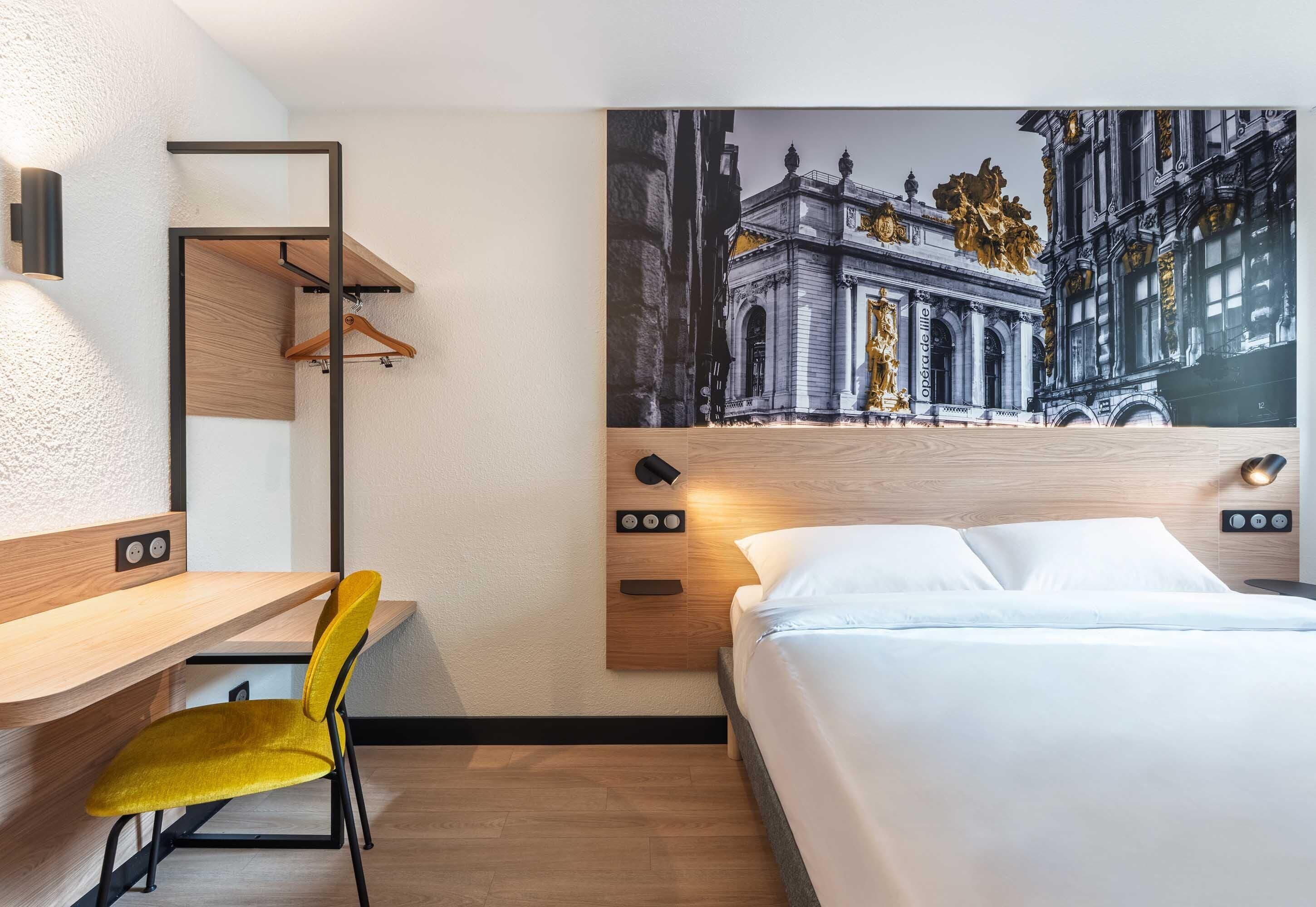 Et hotelværelse med seng, skrivebord, stol, væglampe og et stort muralmaleri af en bygning.