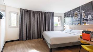A hotel room with a bed, a window with curtains, a chair, and a wall with a cityscape painting.