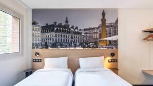 A hotel room with two beds, a cityscape mural on the wall, a wooden headboard, and a small table with a lamp. - B&B Hotel Lille Roubaix Centre Grand-Place (Roubaix)
