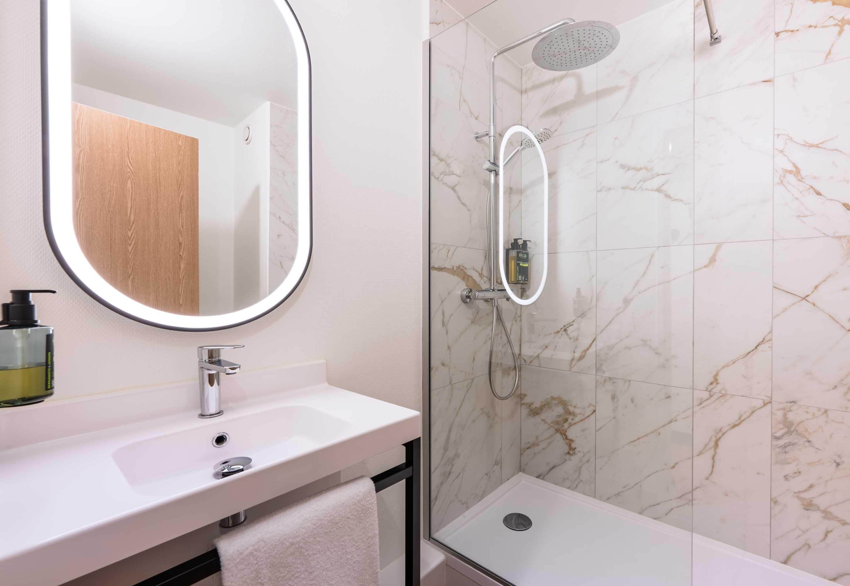 A bathroom with a white sink, a mirror, a shower with a handheld showerhead, and a marble-tiled wall.