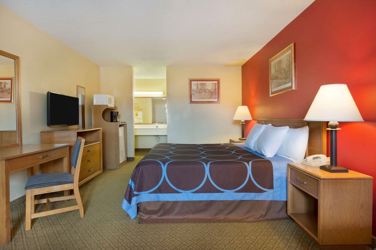 A hotel room with a bed, desk, chair, TV, bedside lamps, and a bathroom visible in the background.