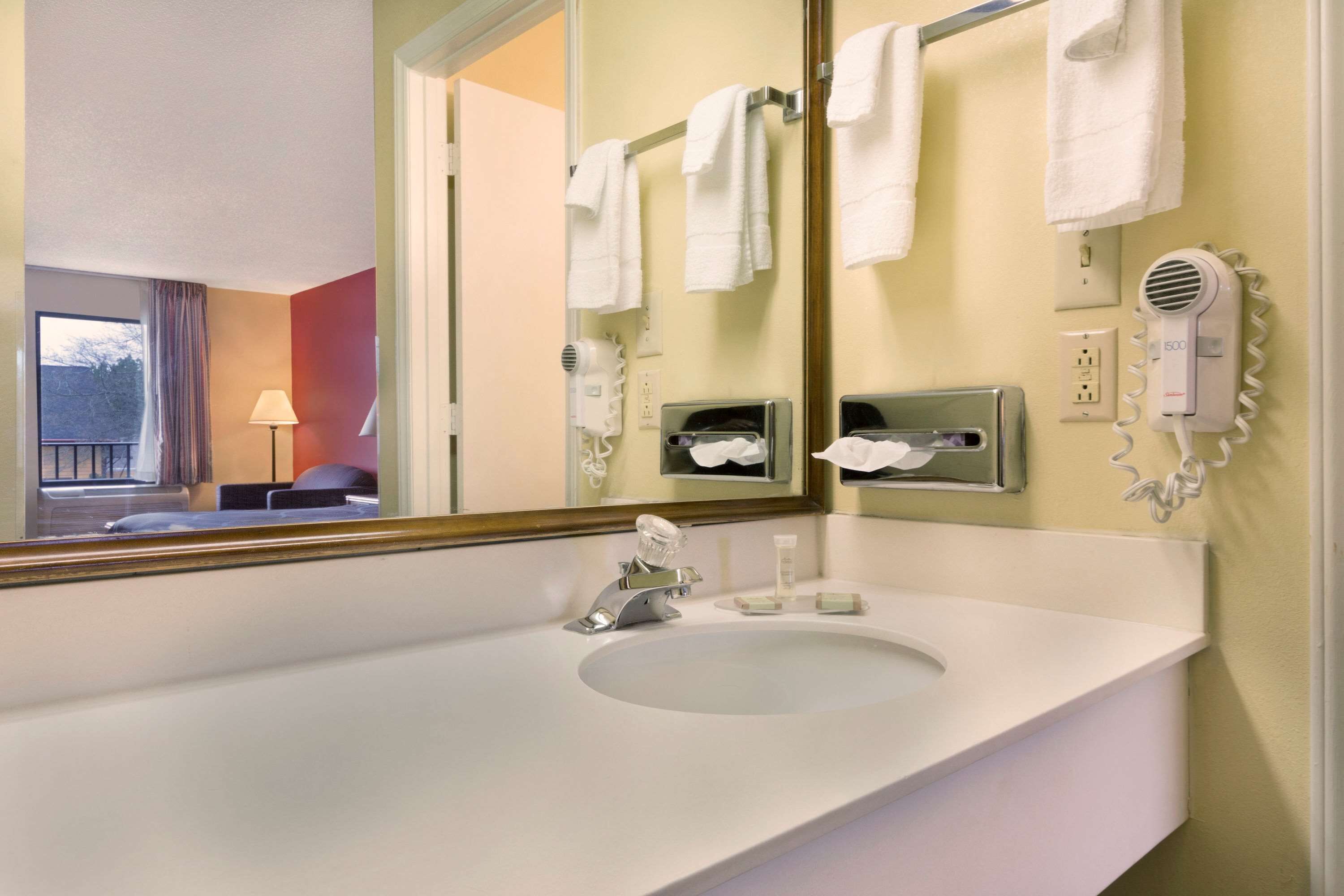 A hotel bathroom with a large mirror, white sink, towel rack, hairdryer, and a window with curtains.