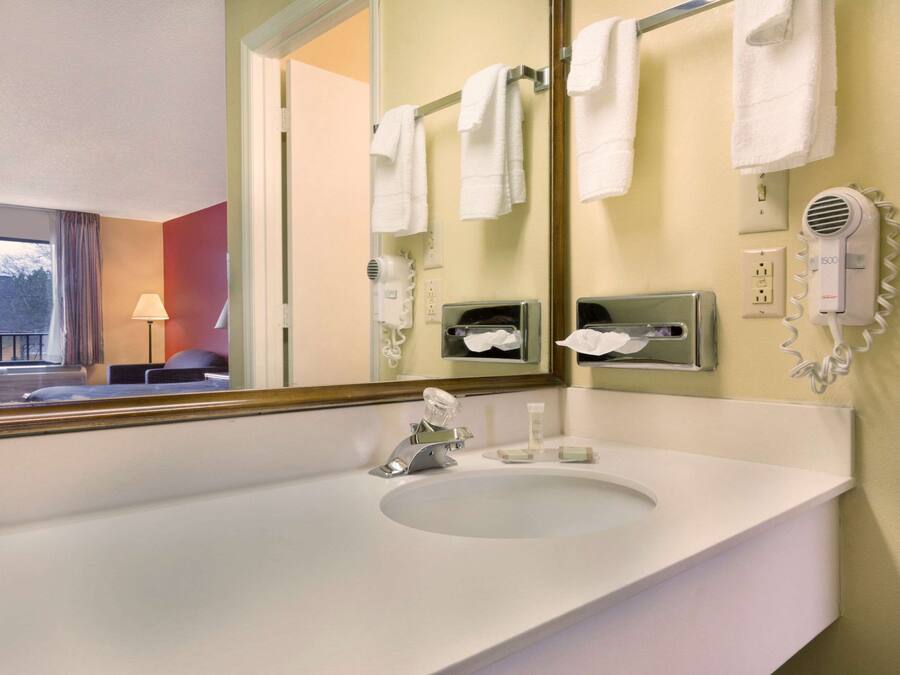 A hotel bathroom with a large mirror, white sink, towel rack, hairdryer, and a window with curtains.