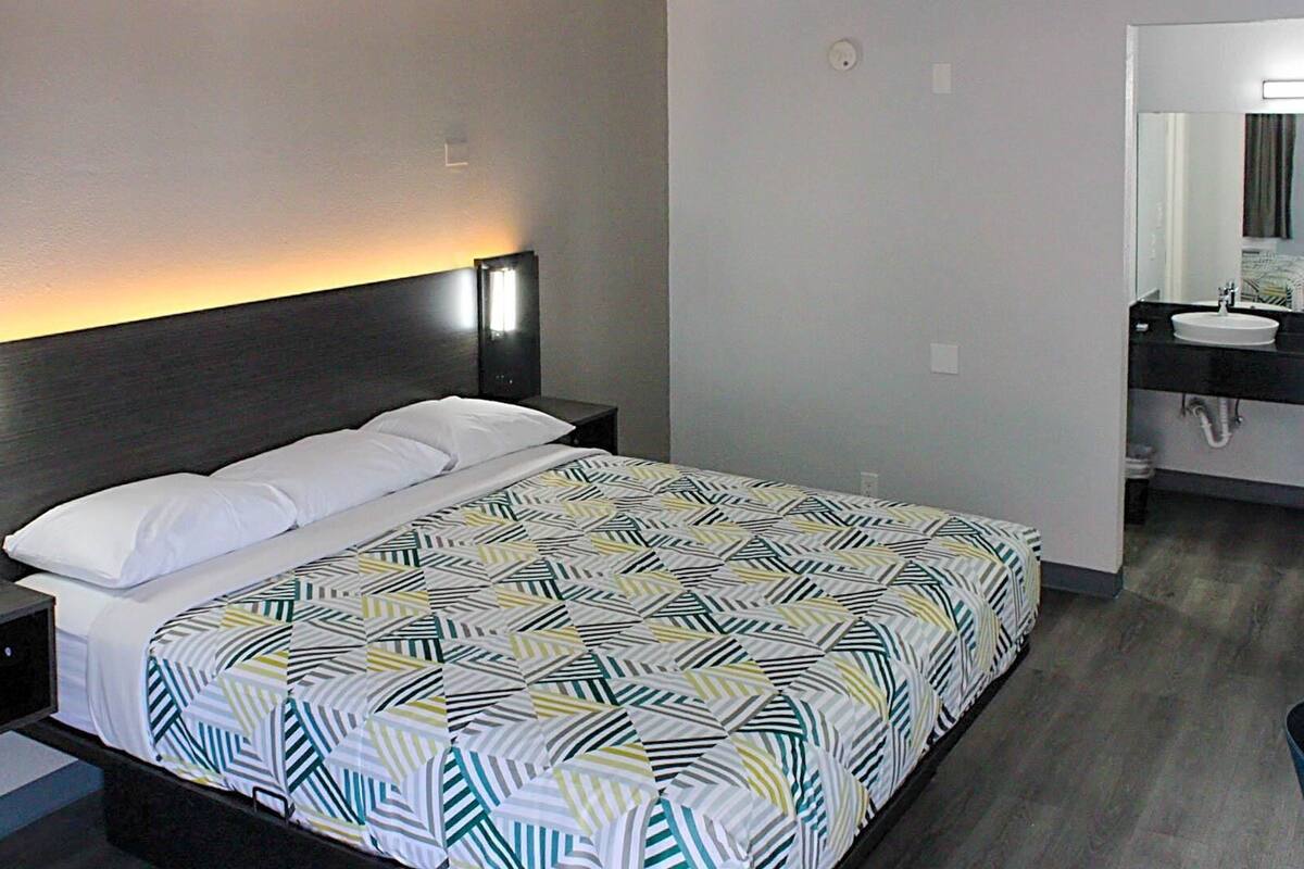 A modern hotel room with a neatly made bed, a bedside table with a lamp, and a bathroom with a sink and mirror.