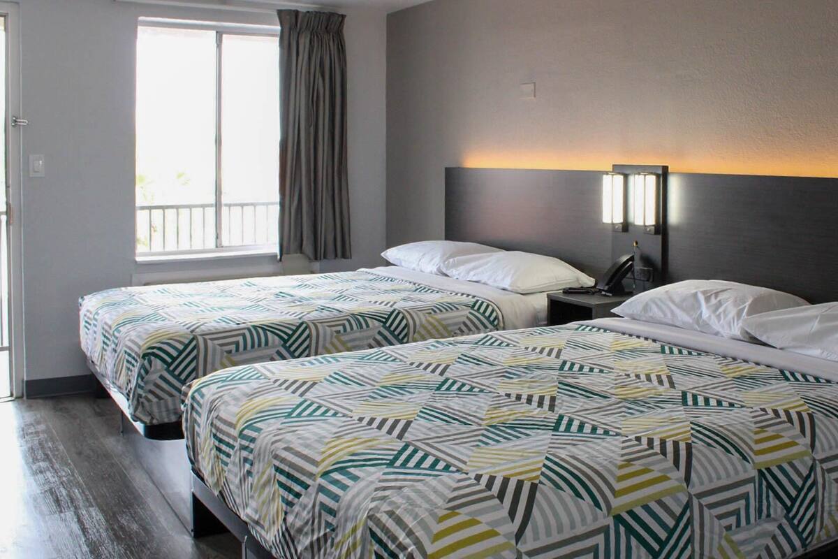 A hotel room with two beds, a nightstand, a lamp, and a window with curtains.