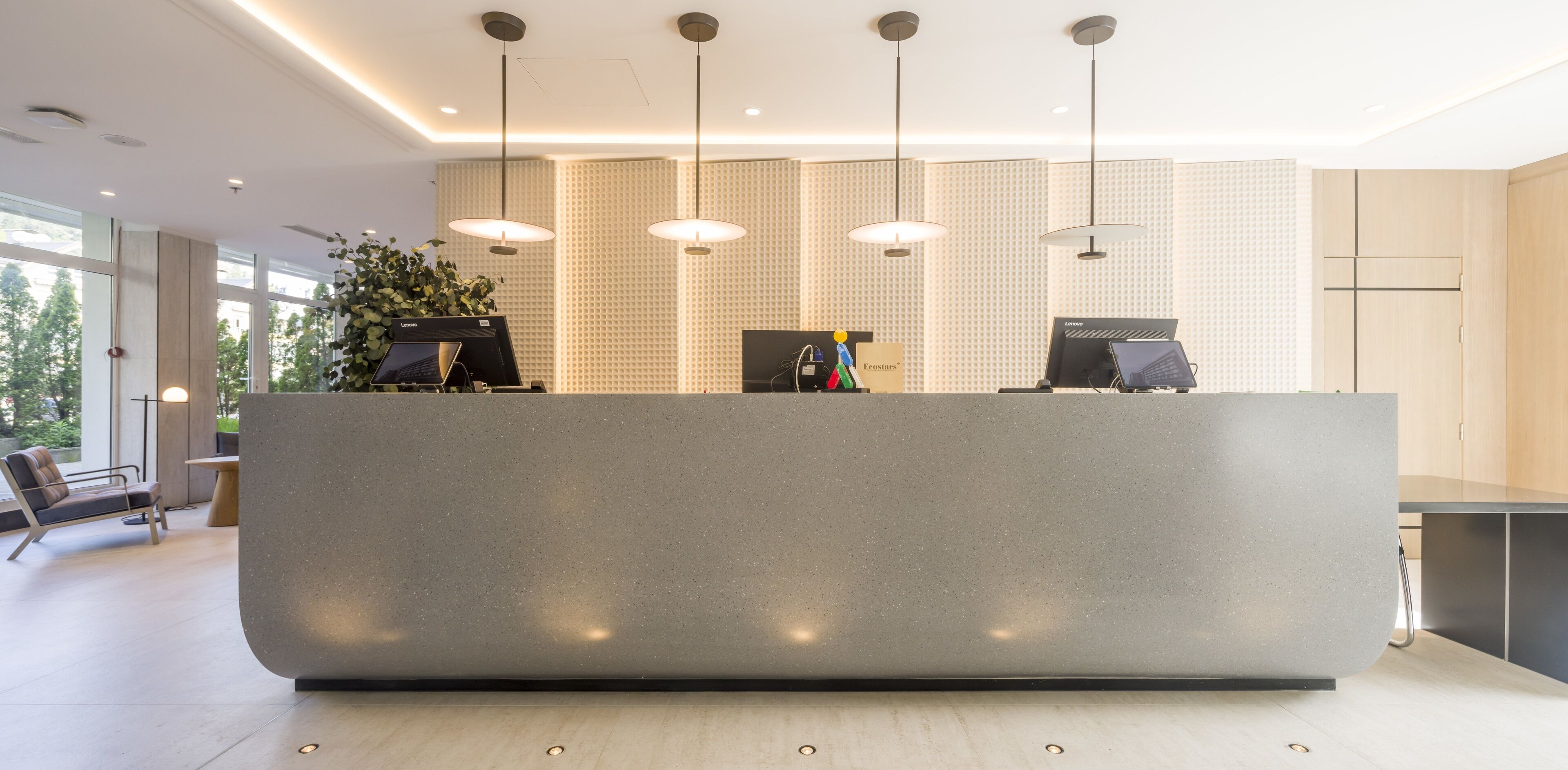 A modern reception desk with two computer monitors, a plant, and a seating area with a bench and a chair.
