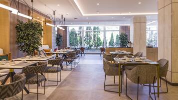 Un comedor moderno con varias mesas y sillas, ventanas amplias con vistas a árboles, y plantas de interior.
