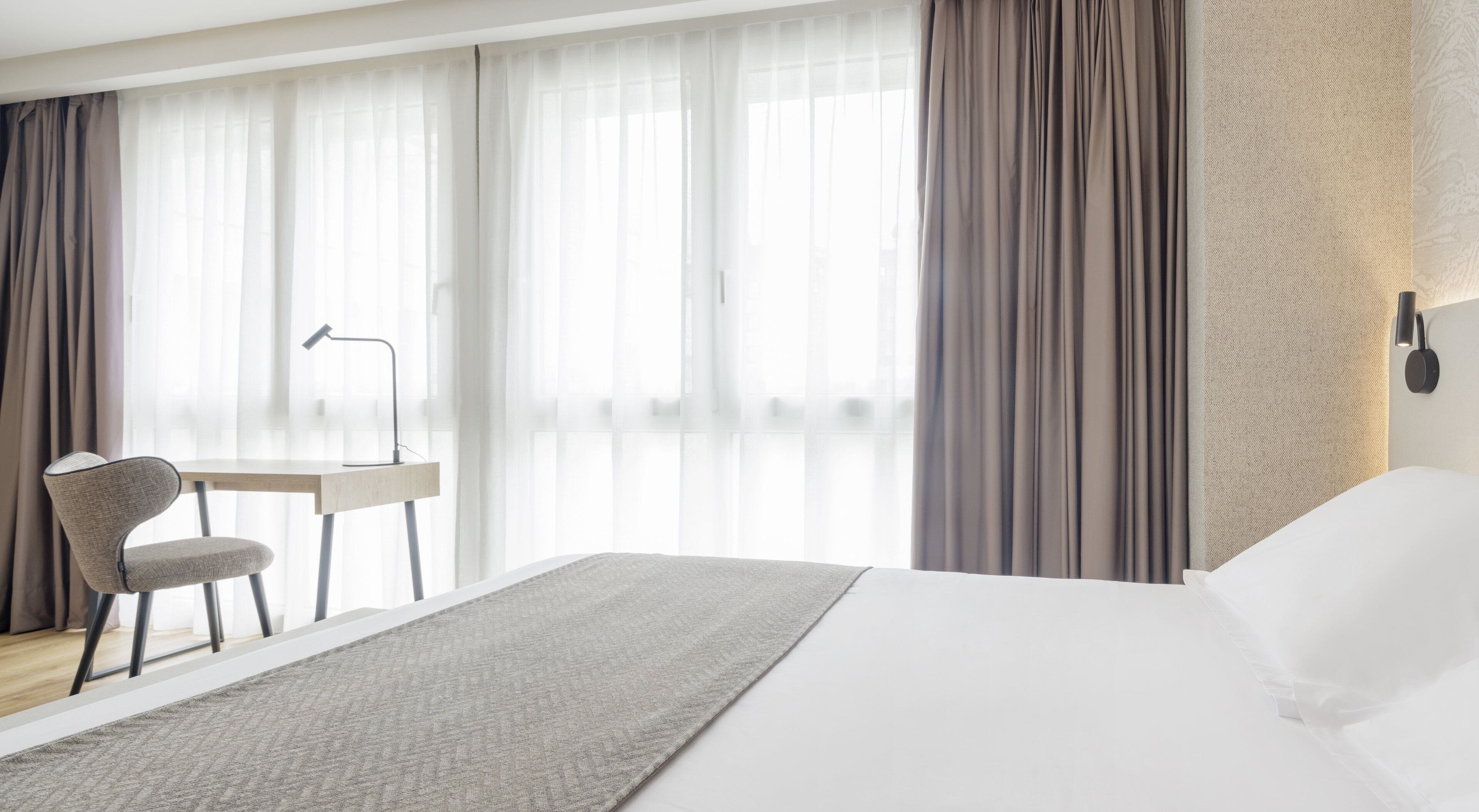 A modern hotel room with a large bed, a desk with a chair, and a lamp.