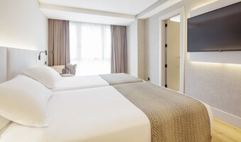A hotel room with two beds, a desk, a chair, a TV, and a large window with curtains.