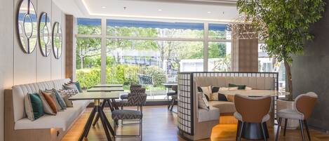 Una sala de estar moderna con sofá, mesa de comedor, sillas y ventanas amplias que ofrecen vistas a zonas verdes y edificios.