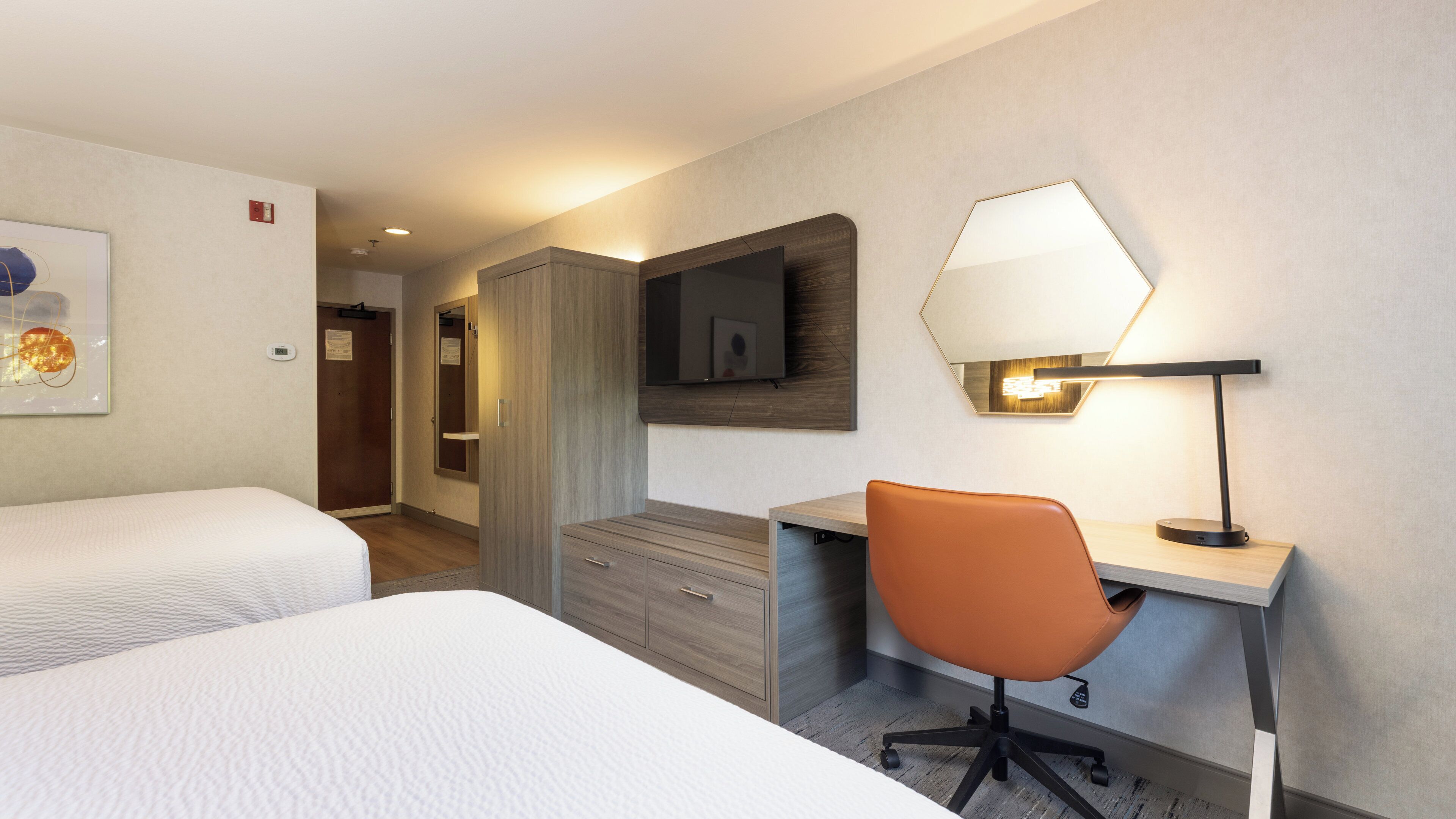 A hotel room with two beds, a desk with a chair, a mirror, and a lamp.