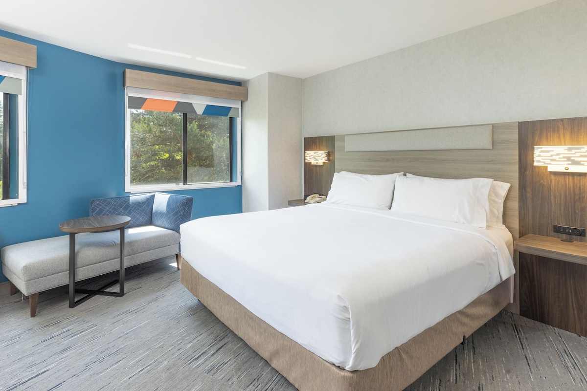 A modern hotel room with a large bed, a seating area, and a view of trees through the windows.