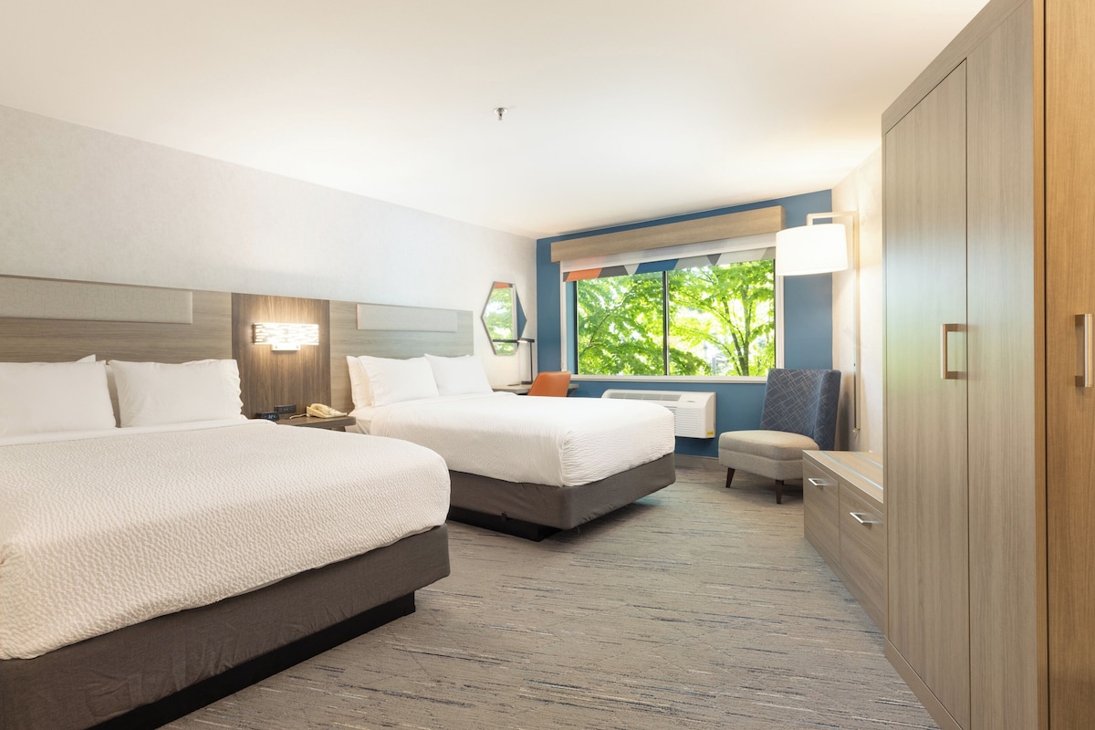 A hotel room with two beds, a desk, a chair, a wardrobe, and a window with a view of trees.