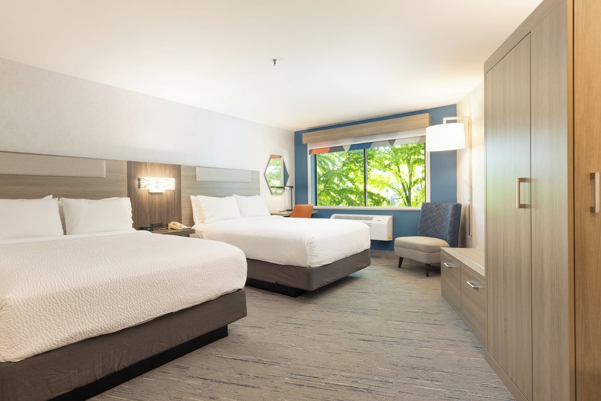 A hotel room with two beds, a desk, a chair, a wardrobe, and a window with a view of trees.
