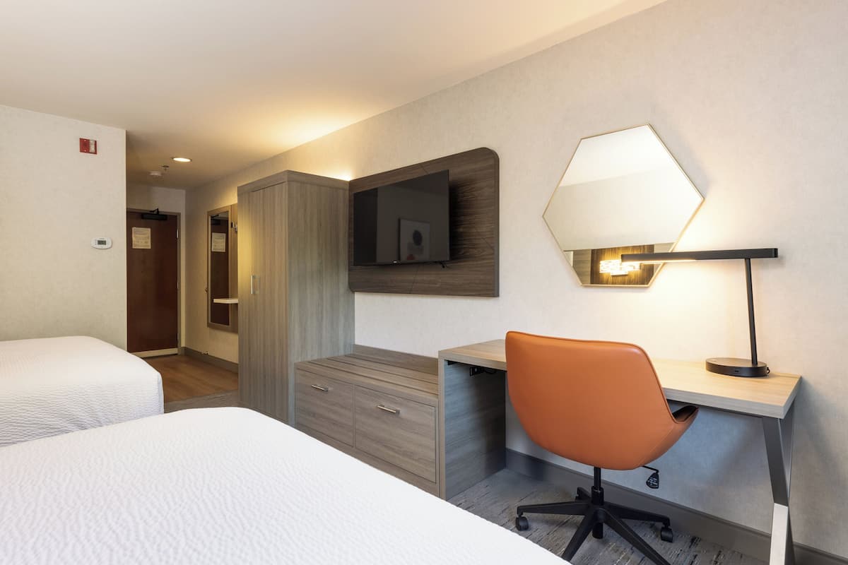 A hotel room with two beds, a desk with a chair, a mirror, and a lamp.