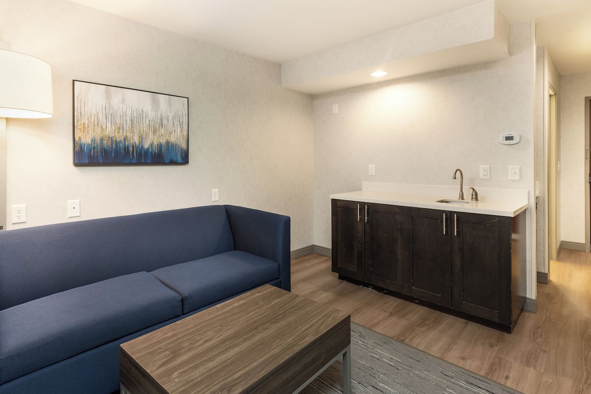 A modern living room with a blue sofa, a wooden coffee table, a kitchen area with dark cabinets, and a painting on the wall.