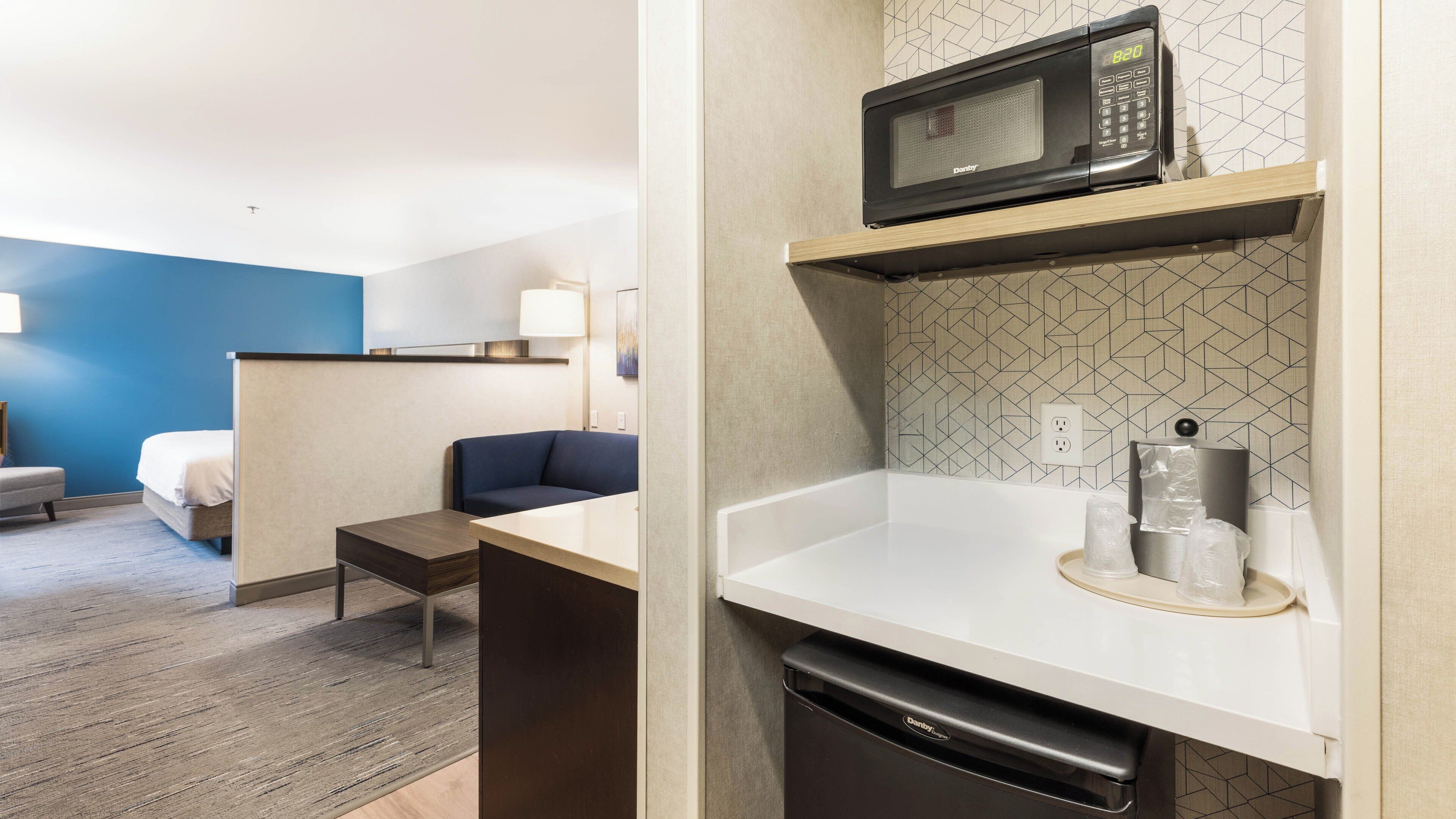 A hotel room with a bed, a desk, a chair, a microwave, and a small kitchen area with a dishwasher.